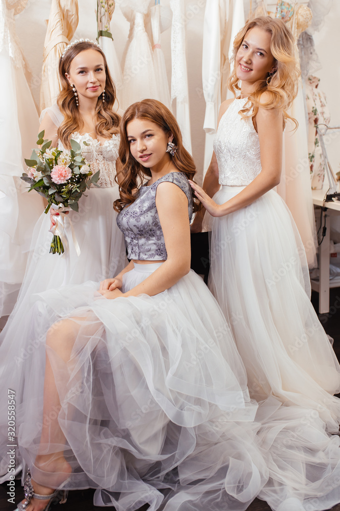 portrait of good-looking three women together in wedding dresses, posing, show new dress made by professional designer tailor, isolated in wedding salon among dresses