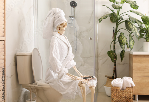 Skeleton in bathrobe with mobile phone sitting on toilet bowl