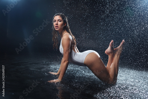 Beautiful sexy wet girl in a white bathing suit sits under raindrops on a black background.