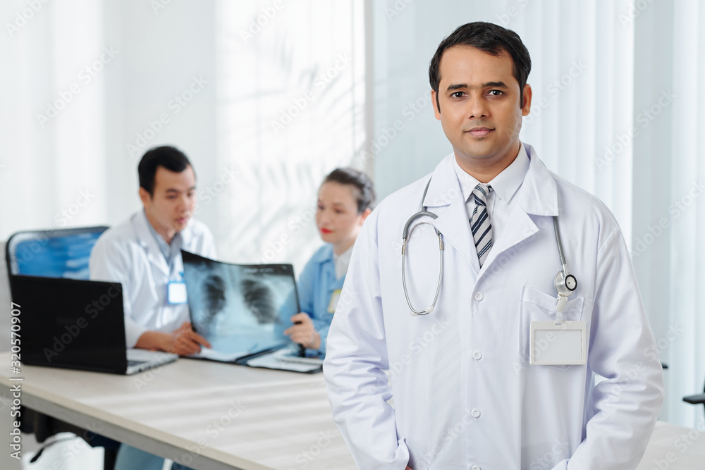 Portrait of confident general practitioner in white labcoat looking at camera, his colleagues discussing x-ray in background Portrait of confident general practitioner in white labcoat looking at camera, his colleagues discussing x-ray in background