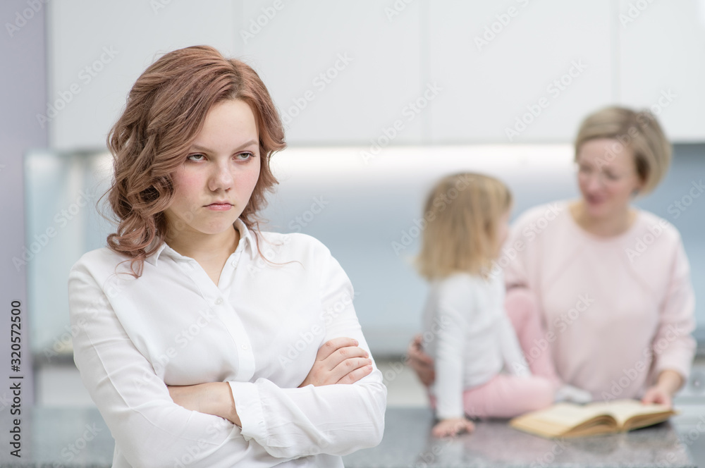 A teenage girl looks upset at the camera, in the background her mother is playing with her younger sister. Problems of puberty, depression, problems in the family