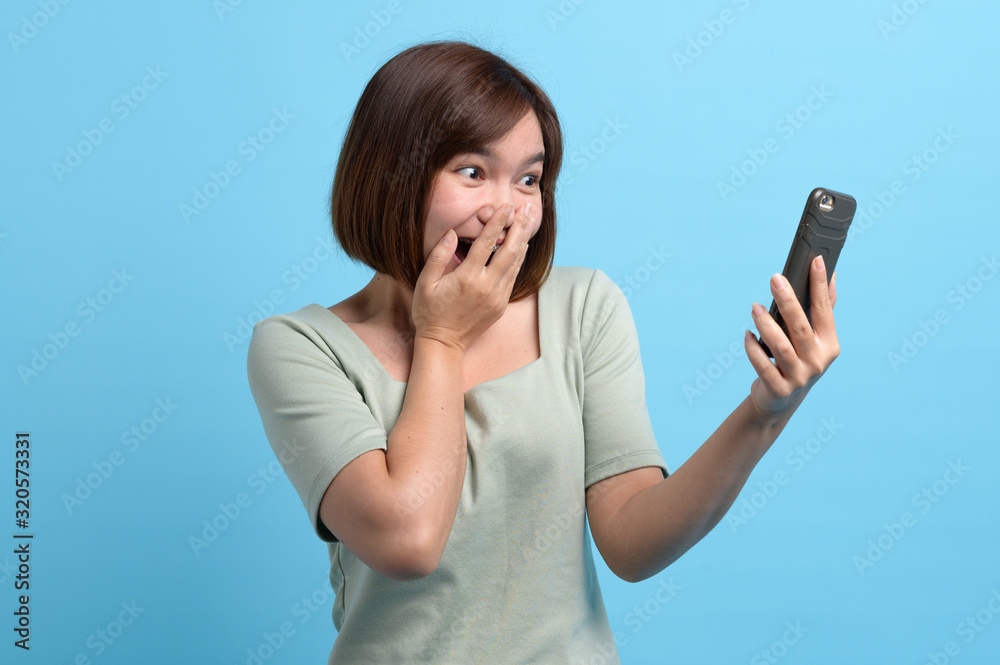 Portrait of shocked asian woman looking in mobile phone and being surprised or astonished to see large discounts sales campaign, isolated over blue background in studio.