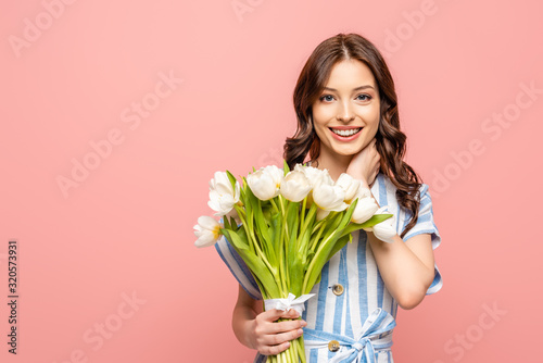happy girl touching neck while holding bouquet of white tulips and smiling at camera isolated on pink