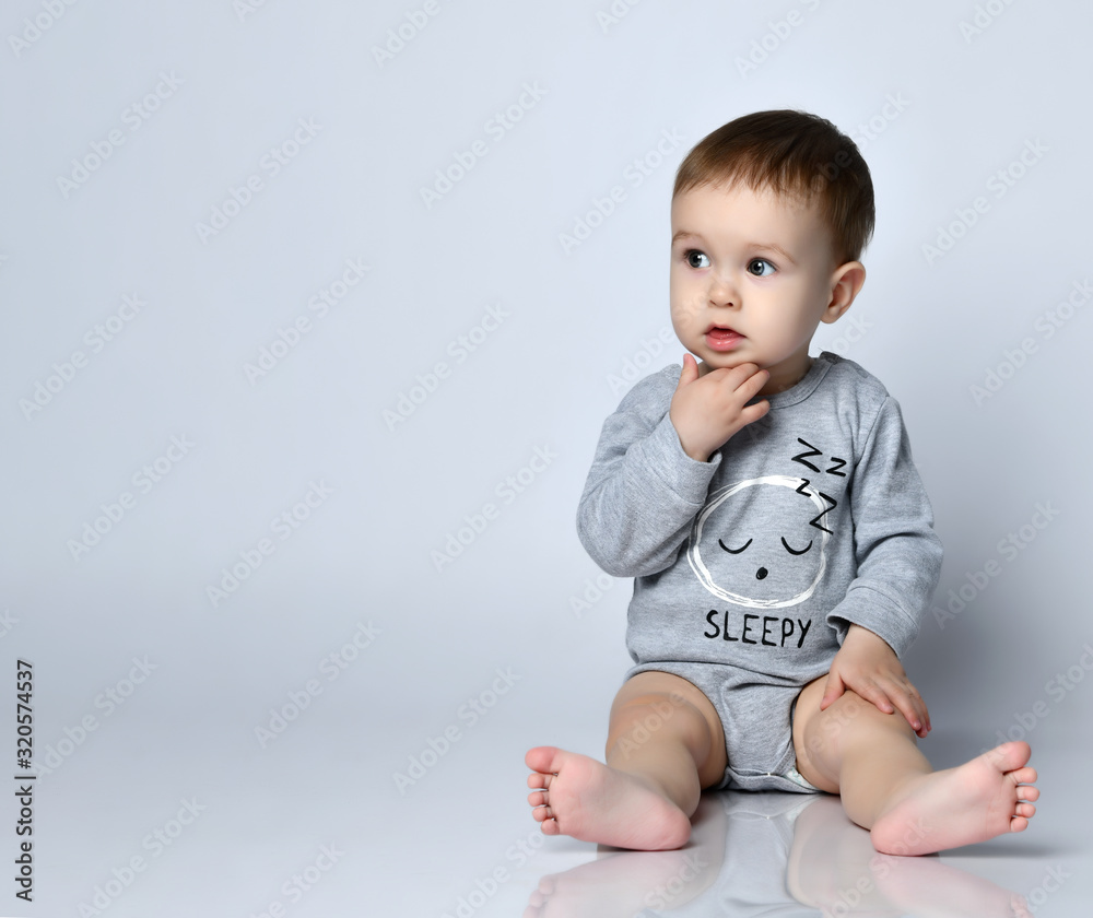 Little baby boy toddler in grey casual jumpsuit, cap and barefoot sitting on floor and feeling interested looking aside Little baby boy toddler in grey casual jumpsuit, cap and barefoot sitting on floor and feeling interested looking aside