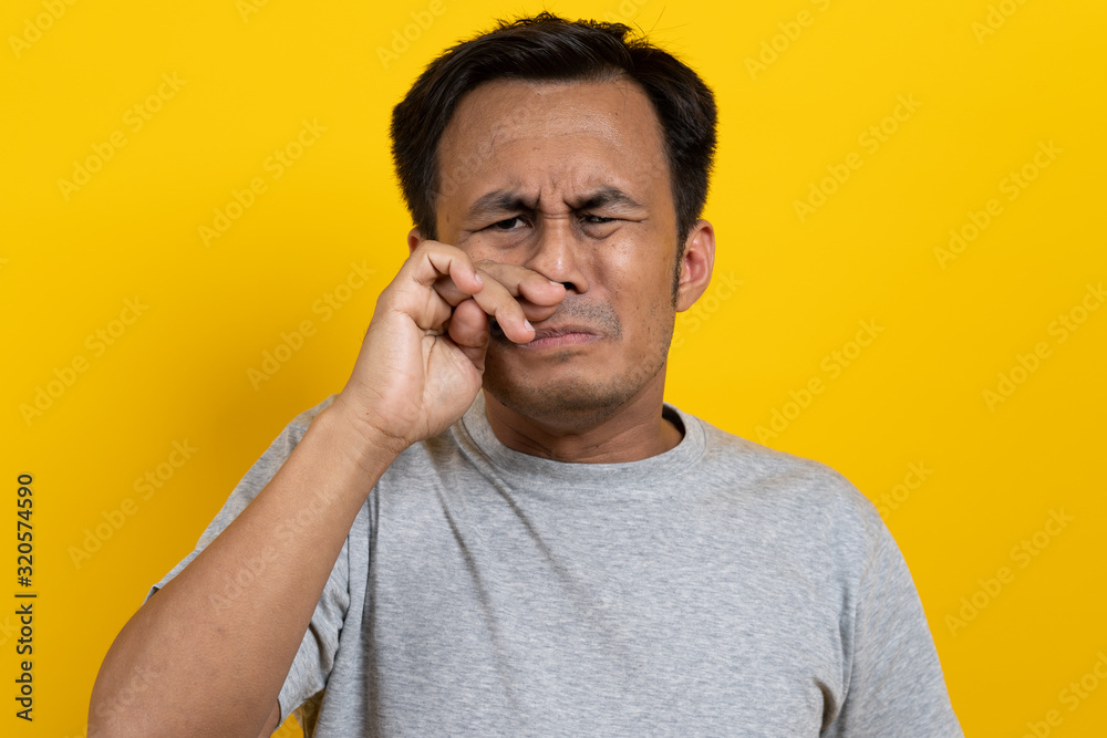 Face expression of human.  Asian male smelling disgusted odor and pinching his nose in studio shoot yellow background.