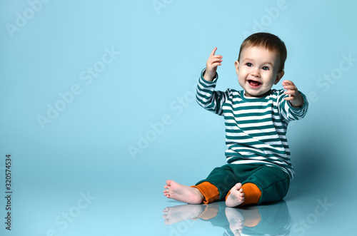 Little baby boy in stylish casual clothing barefoot sitting on floor and smiling with raised hands over blue wall background photo