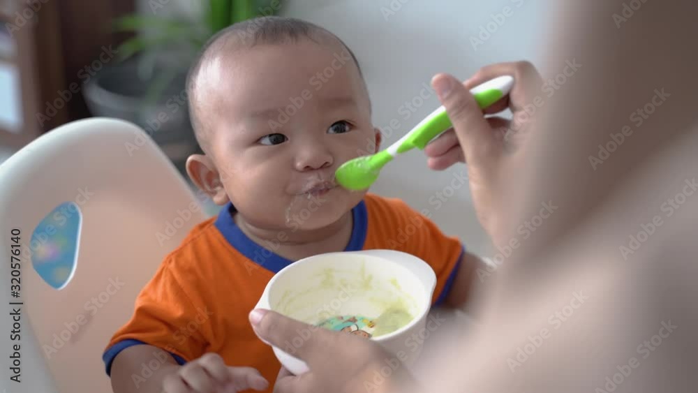 asian Mother feeding her baby son with spoon. Mother giving healthy food to adorable child at home asian Mother feeding her baby son with spoon. Mother giving healthy food to adorable child at home