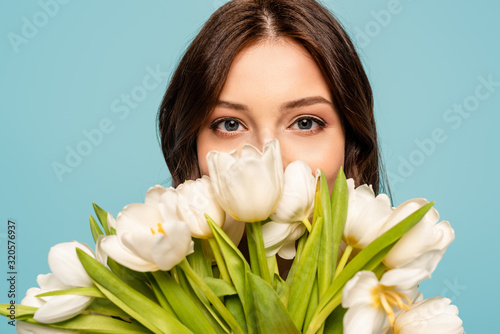 beautiful young woman enjoying flavor of white tulips while looking at camera isolated on blue