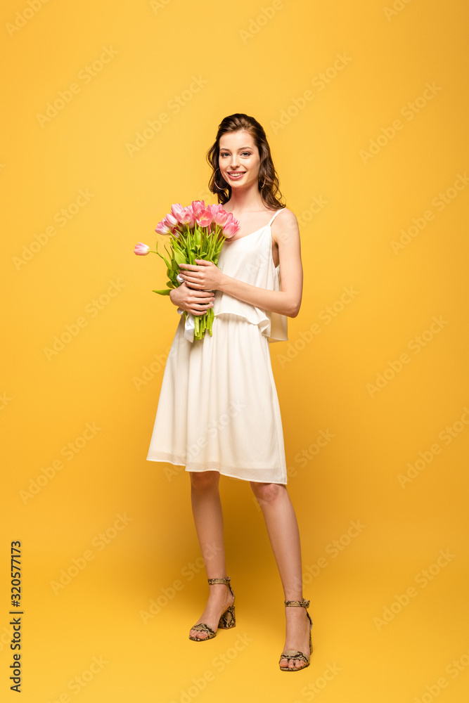 full length view of beautiful young woman smiling at camera while holding bouquet of pink tulips on yellow background full length view of beautiful young woman smiling at camera while holding bouquet of pink tulips on yellow background