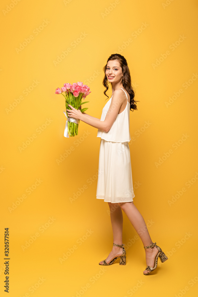 full length view of attractive young woman smiling at camera while holding bouquet of pink tulips on yellow background full length view of attractive young woman smiling at camera while holding bouquet of pink tulips on yellow background