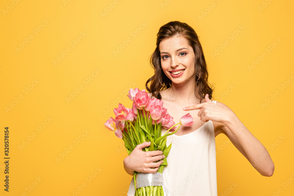 smiling young woman pointing with finger at bouquet of pink tulips while looking at camera isolated on yellow smiling young woman pointing with finger at bouquet of pink tulips while looking at camera isolated on yellow