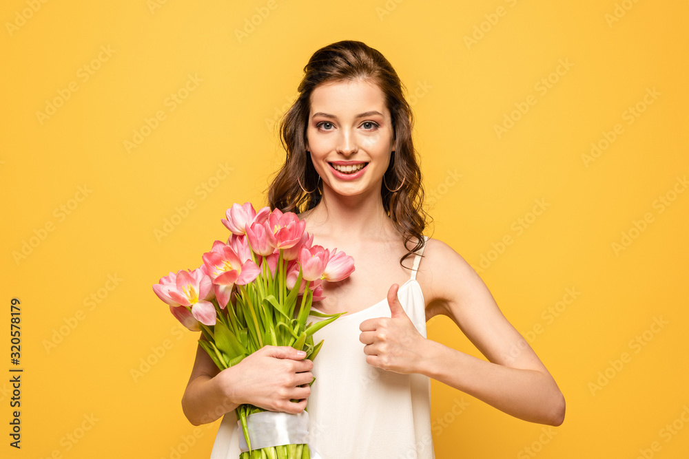 happy young woman holding bouquet of pink tulips while looking at camera and showing thumb up isolated on yellow happy young woman holding bouquet of pink tulips while looking at camera and showing thumb up isolated on yellow