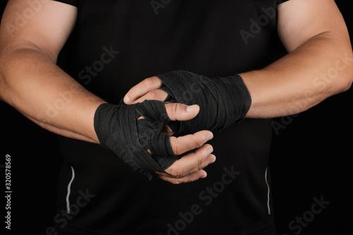 adult athlete stand in black clothes and wrap his hands in black textile elastic bandage