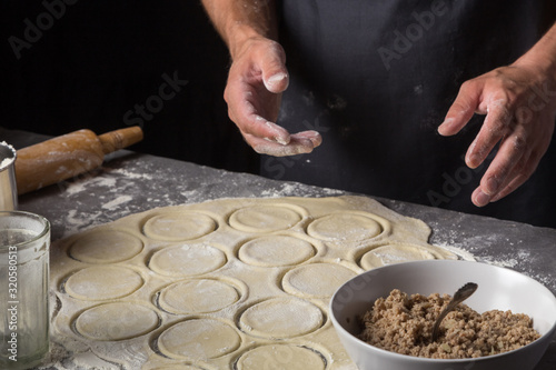 A man is making a dough for varenyky or dumplings, with meat, onions and bacon. Man in a black apron without face. Man squeezes out circles of dough with a glass. Making dough by male hands at bakery 