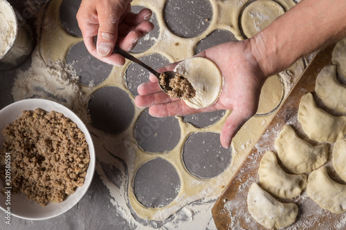 A man is making varenyky or dumplings, with meat, onions and bacon. Man is stuffing the dough with meat. Man in a black apron without face. Making dough by male hands at bakery Stone table close-up ph