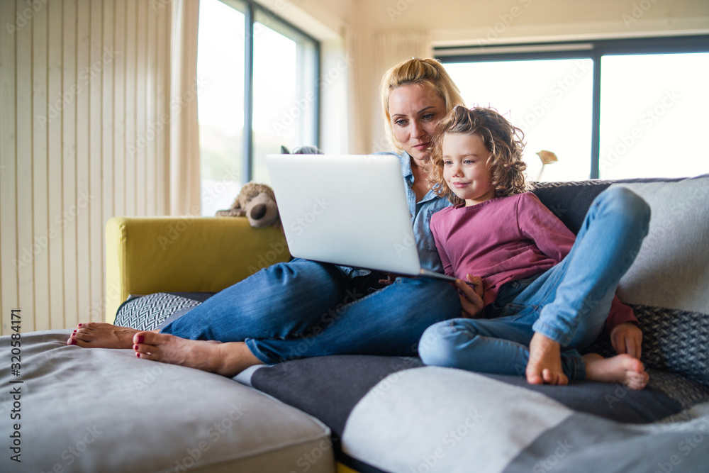 A cute small girl with mother on sofa indoors at home, using laptop.