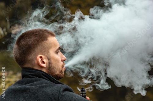 A guy with a beard sits on the lake and smokes an electronic cigarette. Blows puffs of smoke. The concept of unity with nature. Rest near the water. Vape, Man Vaping. lips and smoke, a lot of smoke. photo