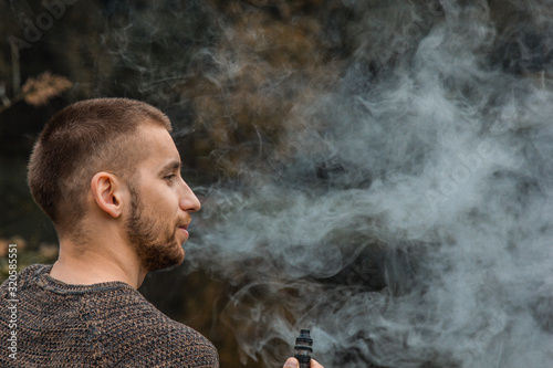 A guy with a beard sits on the lake and smokes an electronic cigarette. Blows puffs of smoke. The concept of unity with nature. Rest near the water. Vape, Man Vaping. lips and smoke, a lot of smoke.