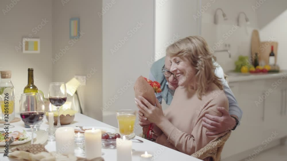 Romantic senior man giving bouquet of flowers to happy wife and embracing her before having holiday dinner at home Romantic senior man giving bouquet of flowers to happy wife and embracing her before having holiday dinner at home