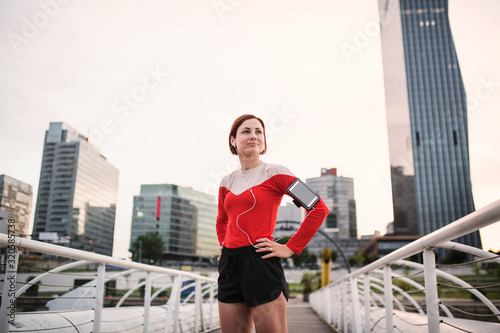 Young woman with smartphone standing outdoors in city, resting after exercise.