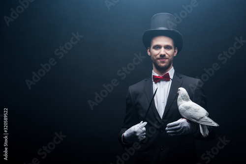 Fotobehang smiling magician in hat showing trick with dove and wand in dark room with smoke