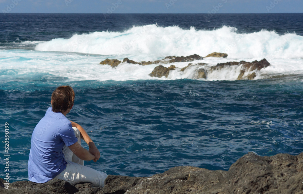 Young man sitting on rock shore and looking at ocean. View from behind. Blue ocean with high waves, water splash