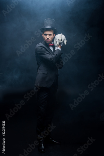 serious magician in suit and hat holding white rabbit, dark room with smoke