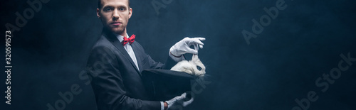 Fotografering panoramic shot of young magician in suit showing trick with white rabbit in hat,