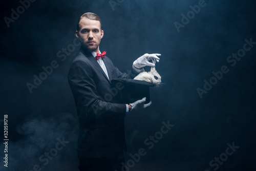 Canvas professional magician in suit showing trick with white rabbit in hat, dark room