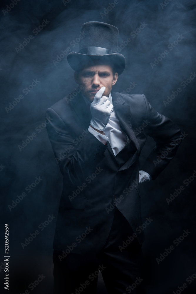 thoughtful magician in suit and hat standing in dark smoky room thoughtful magician in suit and hat standing in dark smoky room