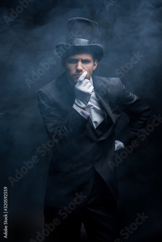 thoughtful magician in suit and hat standing in dark smoky room