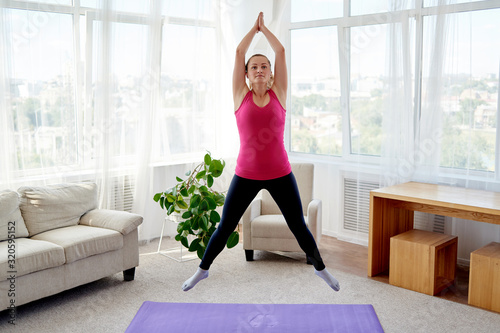 Young fitness woman doing jumping jacks or star jump exercise at home, copy space. Girl working out, full length portrait. Healthy lifestyle concept photo