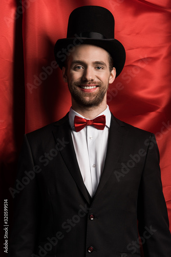 handsome smiling magician in suit and hat in circus with red curtains