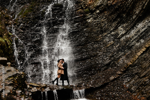young beautiful couple kisses on the background of a falling waterfall  stylish clothes  a man s coat and hat  women s clothing 