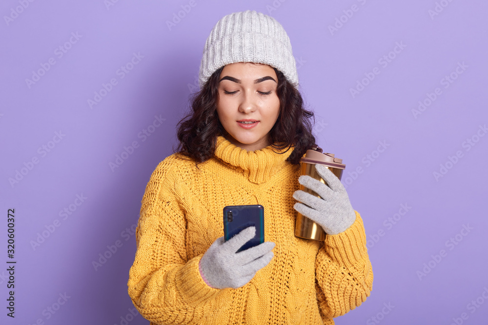 Φωτογραφία Image of good looking adorable attentive female wearing grey hat and gloves, holding smartphone and thermo mug with hot drink in both hands, looking at screen attentively