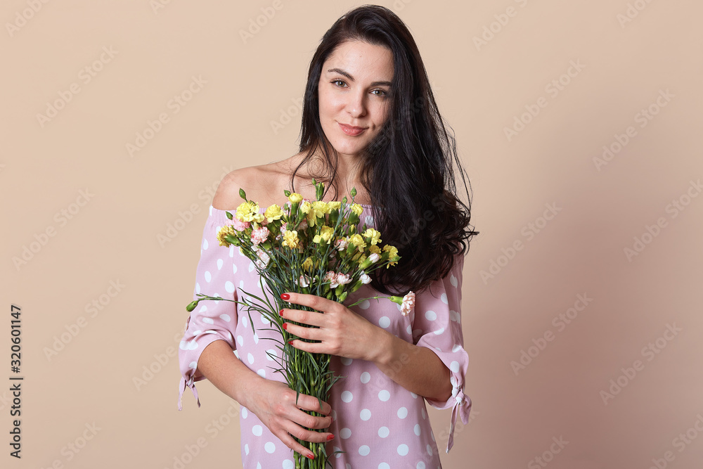 Studio portrait of miling Caucasian woman wearing pink dress, holding bouquet of flowers isolated over beige background, darkhaired female looking at camera. St. Valentines Day and present concept. Studio portrait of miling Caucasian woman wearing pink dress, holding bouquet of flowers isolated over beige background, darkhaired female looking at camera. St. Valentines Day and present concept.