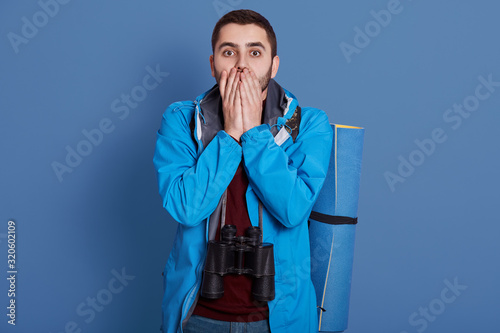 Image of impressed confused handsome explorer covering his mouth with both hands, looking directly at camera, wearing blue jacket, dark blue sweatshirt, having all equipment for hiking alone. photo