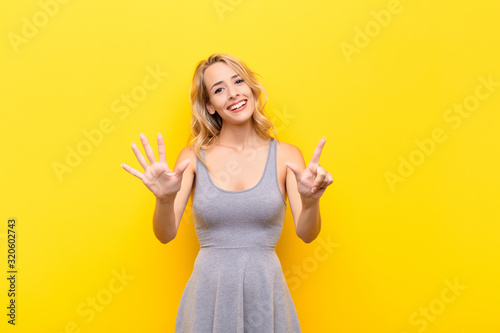 young blonde woman smiling and looking friendly, showing number seven or seventh with hand forward, counting down against orange wall