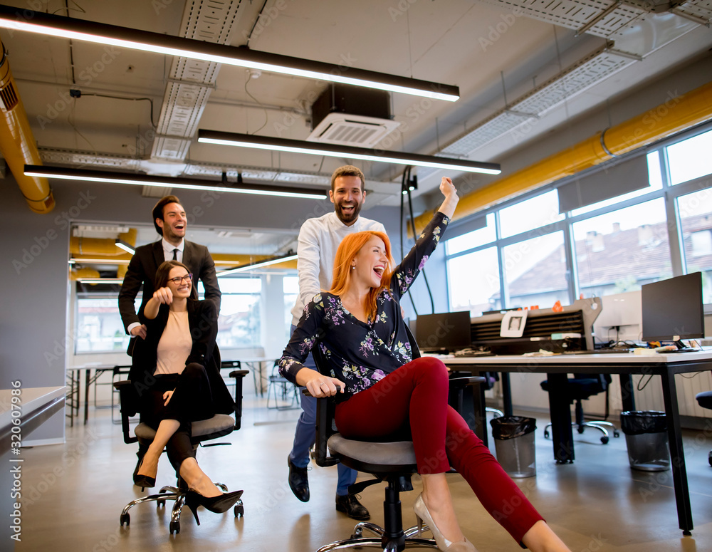 Fototapeta premium Young business people in smart casual wear having fun while racing on office chairs and smiling
