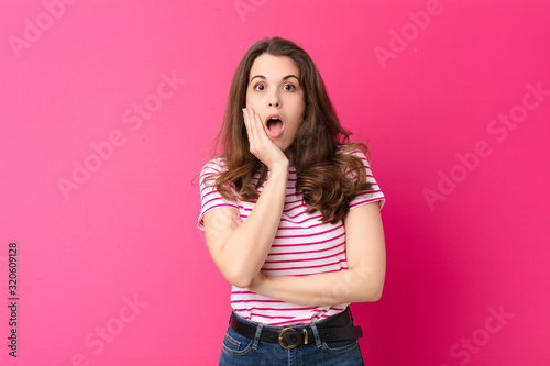 young pretty woman open-mouthed in shock and disbelief, with hand on cheek and arm crossed, feeling stupefied and amazed against pink wall photo