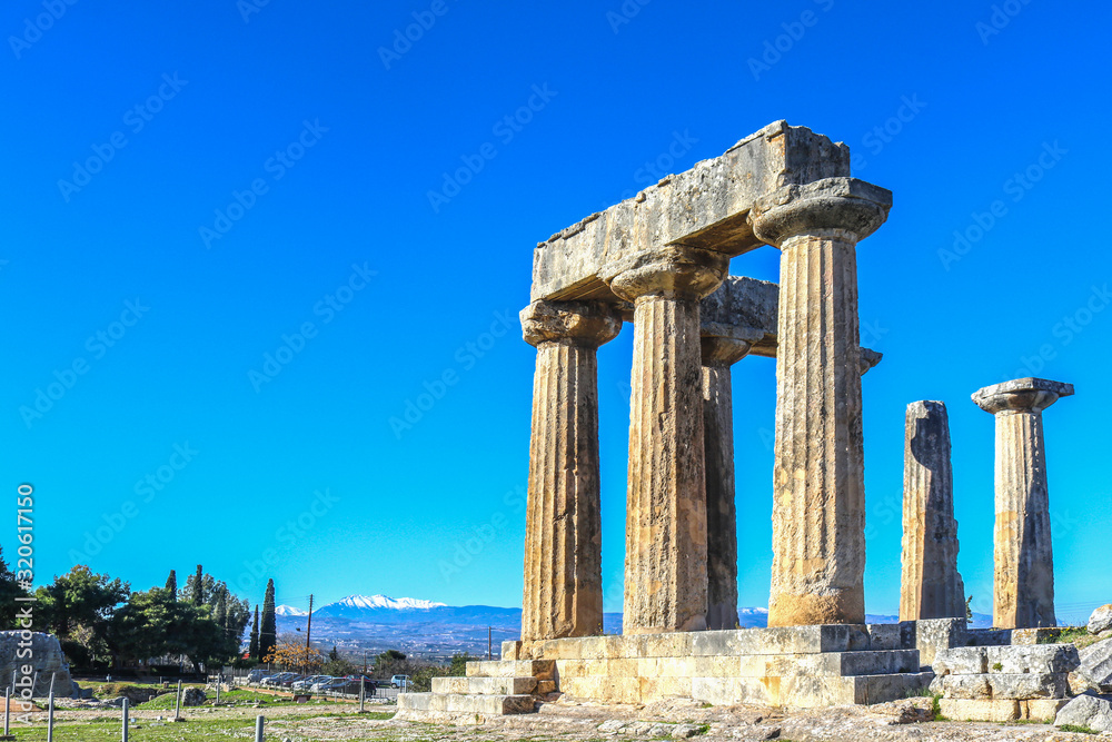 Fototapeta premium Ruins and columns at Ancient Corinth Greece under extremely blue sky with snow-capped mountains of the mainland across the Corinth Canal in the distance