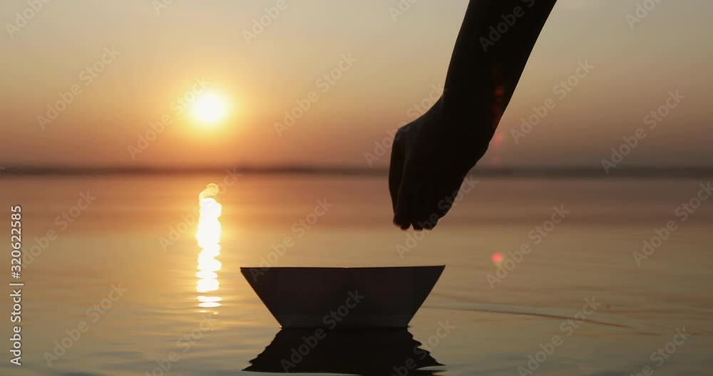 Silhouette of a human hand letting go a paper boat in water of the sea or lake at sunset. T