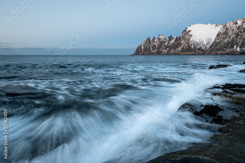 Waves in winter landscape crushing on dramtic rocks with snow covered Oksen mountains in background, Tungeneset, Ersfjord, Senja, Norway
 photo