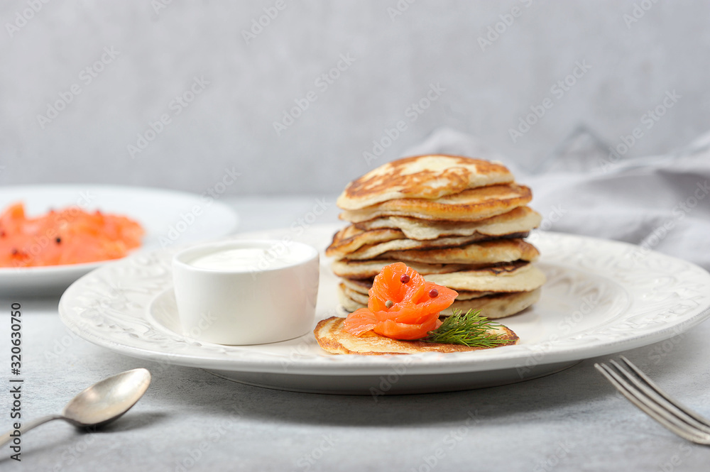 Pancakes with salted salmon. Fritters served with salmon and sour cream. Cutlery and a napkin complete the table. Light background. Close-up. Pancakes with salted salmon. Fritters served with salmon and sour cream. Cutlery and a napkin complete the table. Light background. Close-up.