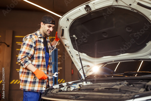 young workman holding wrench standing at the car with open hood and looking at engine