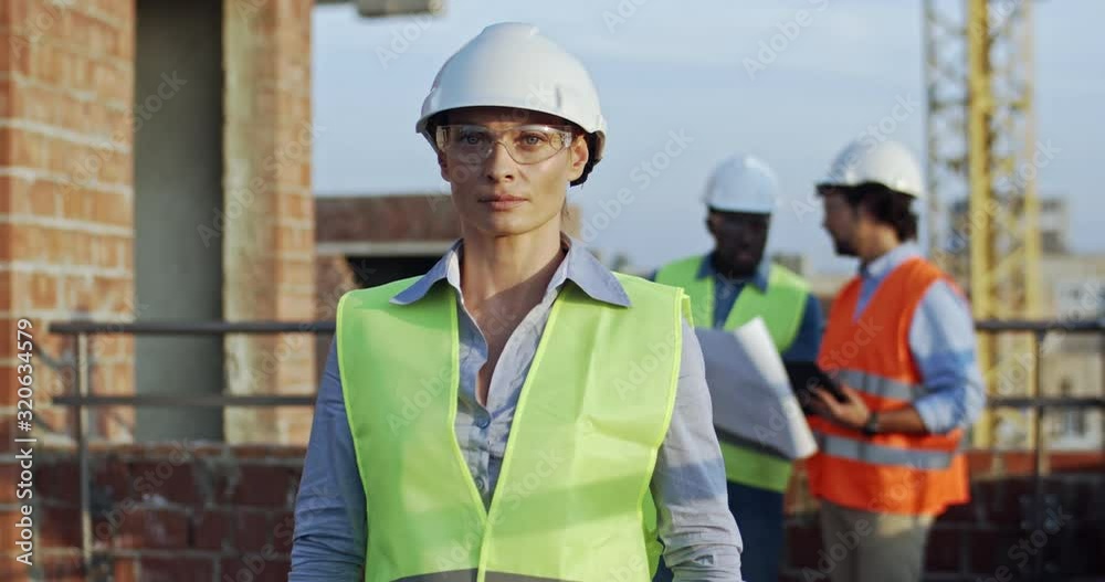 Portrait of the beautiful Caucasian blond woman in hardhat and goggles smiling and posing to the camera at the building site. Two multiethnic men talking about work on the background. Portrait of the beautiful Caucasian blond woman in hardhat and goggles smiling and posing to the camera at the building site. Two multiethnic men talking about work on the background.