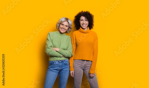 Portrait of two happy lovely sisters one with blonde hair another with curly hair looking at camera laughing isolated on yellow studio wall.
