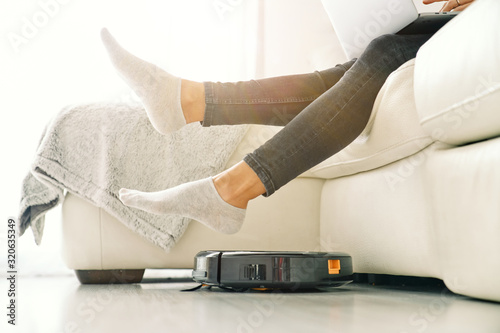 Side view of crop female in jeans and socks sitting on white sofa in room with laminate floor and putting feet on robotic vacuum cleaner photo