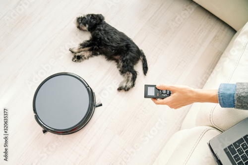 From above crop hand of female using remote control for robotic vacuum cleaner while sitting on sofa with laptop and little dog sleeping on light wooden floor photo