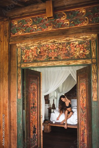 Slim tan woman waking up and enjoying morning coffee in traditional wooden asian bed with white canopy. Wearing black lingerie. View through door of traditional balinese house. Bali vacations. 